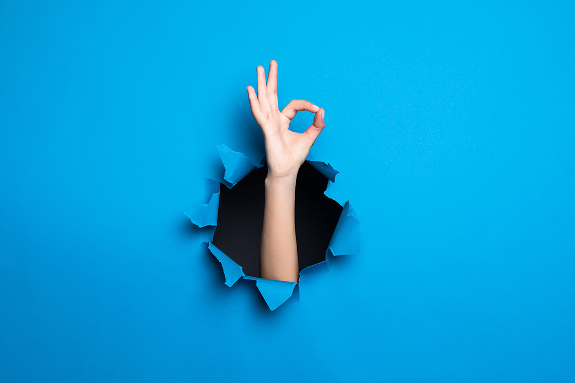 close-up-woman-hand-with-okay-gesture-through-blue-hole-paper-wall-botkeeper