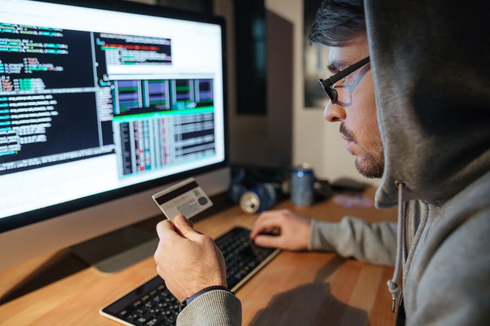 Concentrated young hacker in glasses stealing money from different credit cards sitting in dark room | Botkeeper