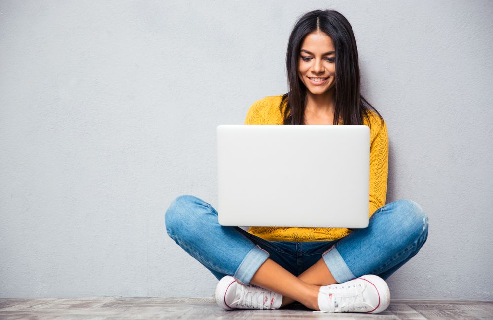 Happy young woman sitting on the floor with crossed legs and using laptop on gray background-1