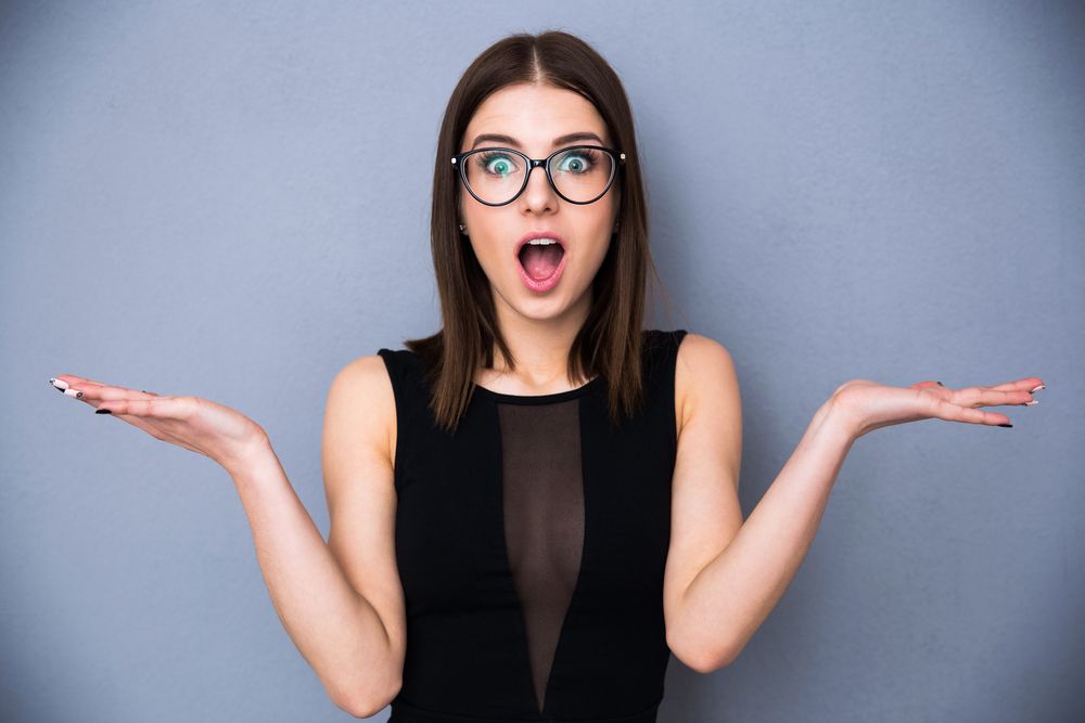 Young beautiful woman with facial expression of surprise standing over gray background. Wearing in trendy black dress and glasses. Looking at the camera | Botkeeper