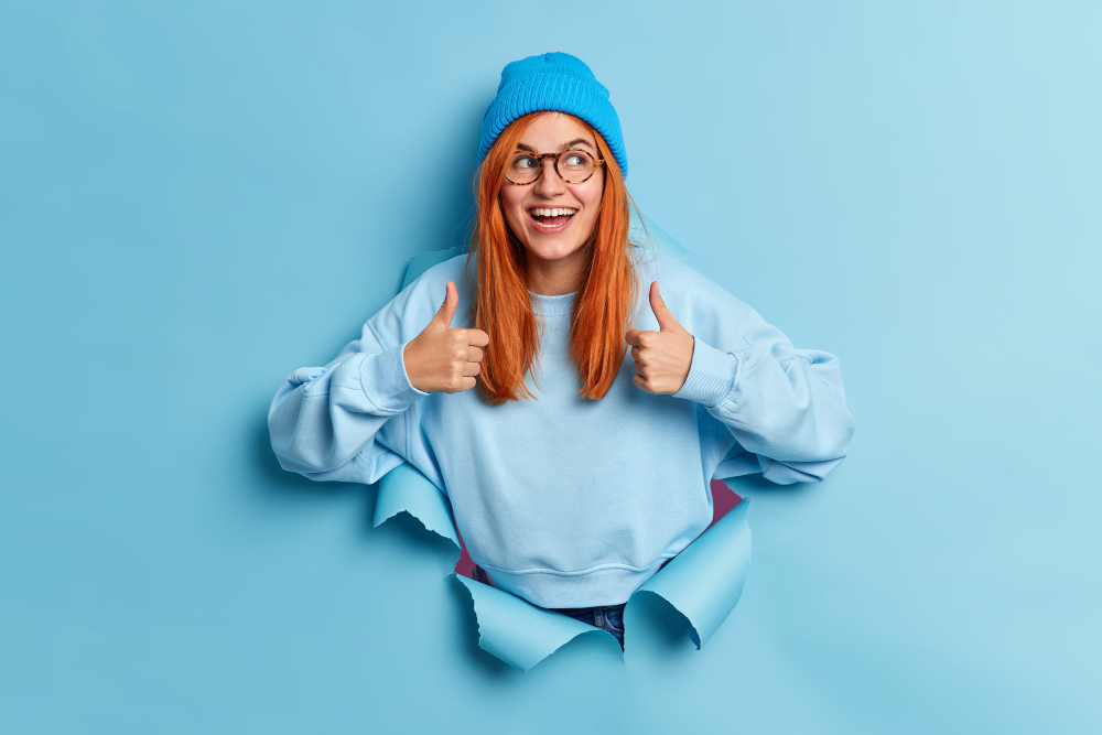 positive-millennial-girl-with-natural-ginger-hair-recommends-sales-keeps-thumbs-up-smiles-gladfully-gives-approval-wears-blue-hat-sweatshirt-breaks-through-paper-hole