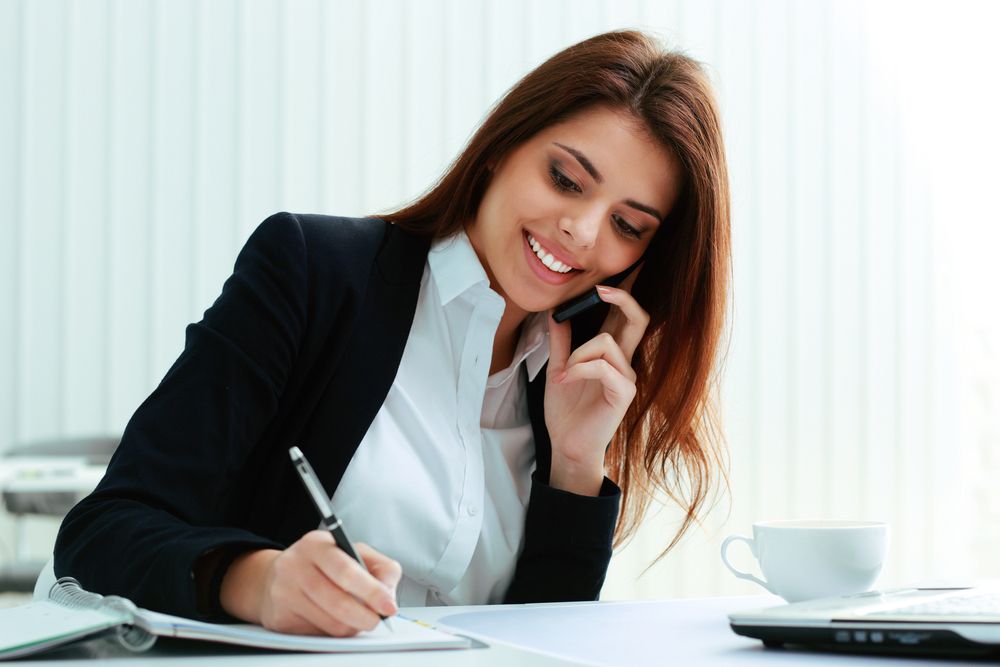 Young happy businesswoman talking on the phone and writing notes in office